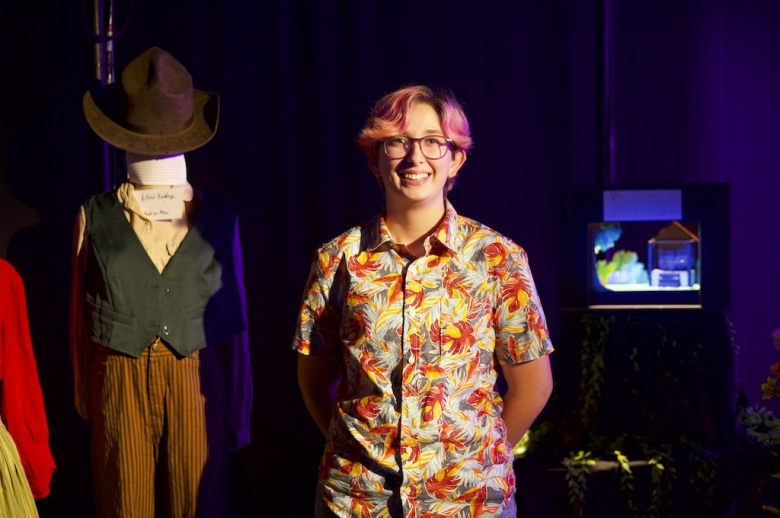 A student in a floral shirt smiles and poses beside a mannequin wearing a dark vest and orange striped trousers, part of the display of their final costume design project.